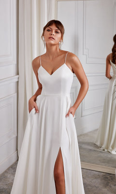 A woman wearing the Aerie Couture 2001 long white dress with thin straps, corset back, and thigh-high slit stands indoors by a mirror and white curtains.