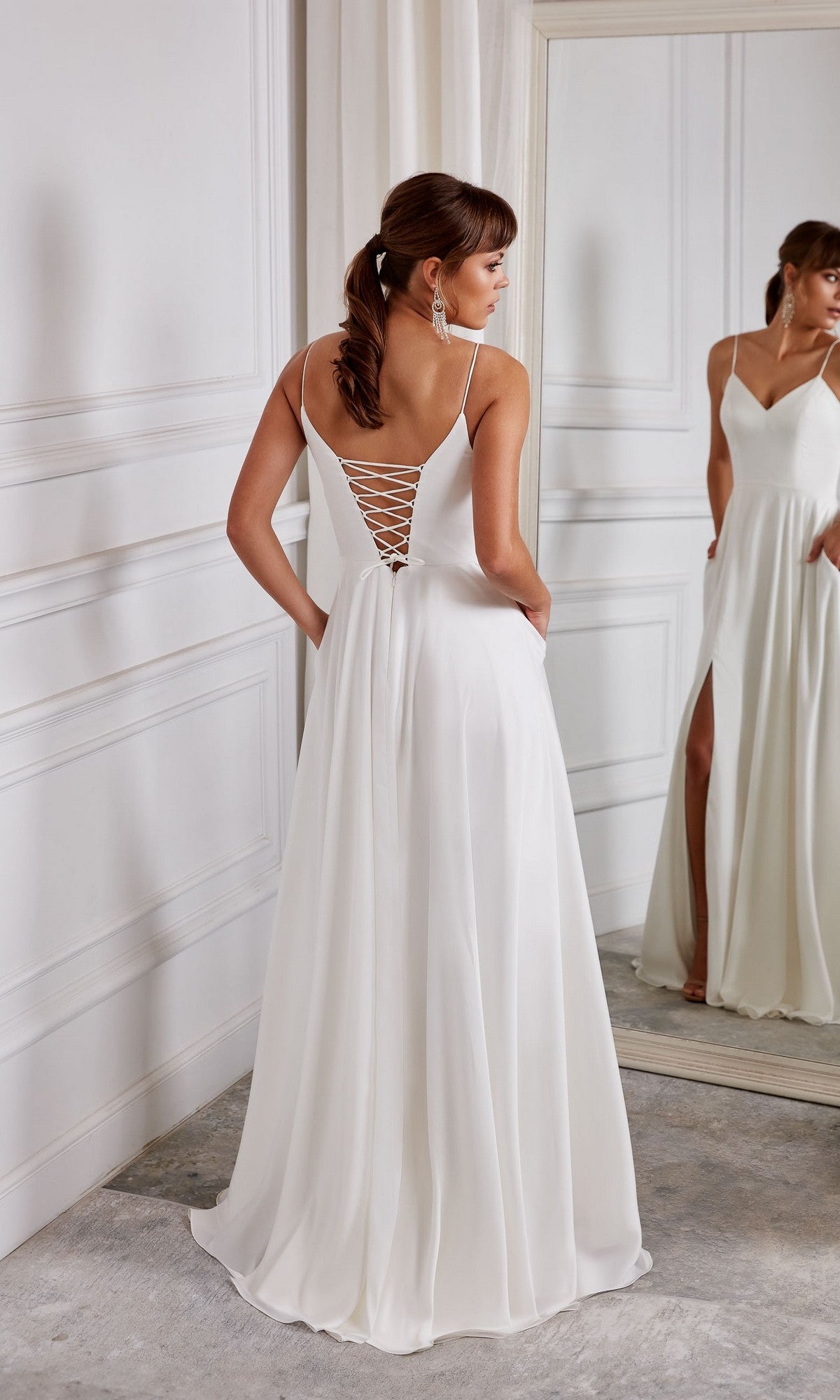 A woman stands in front of a mirror in a white room wearing the Aerie Couture 2001 long white dress, featuring a corset back, side slit, and an elegant flowy skirt.
