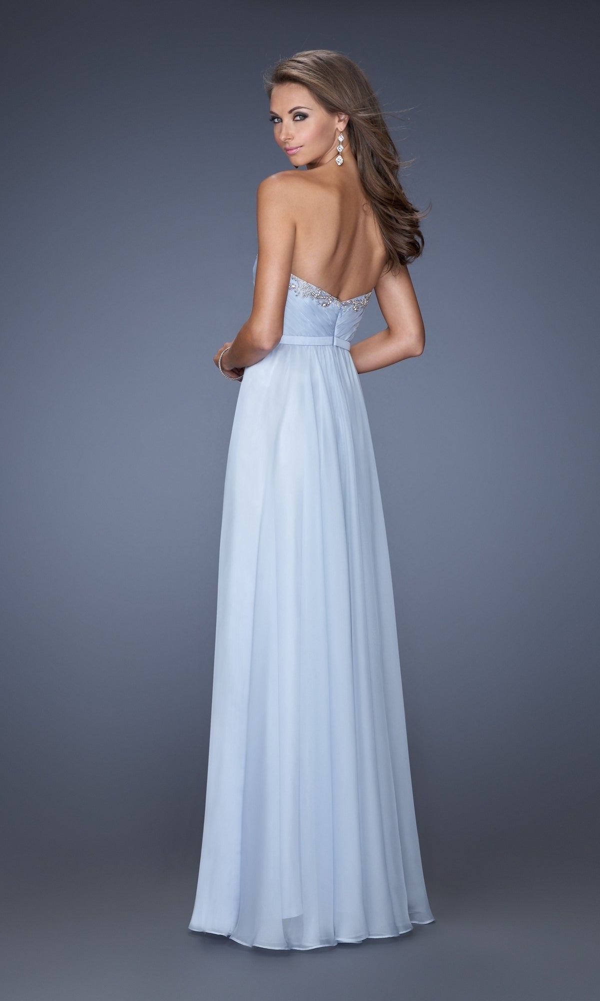 A woman stands against a gray background wearing the La Femme Designer Pleated Chiffon Prom Dress 20027—strapless with a sweetheart neckline, fitted bodice, and flowing floor-length light blue skirt.