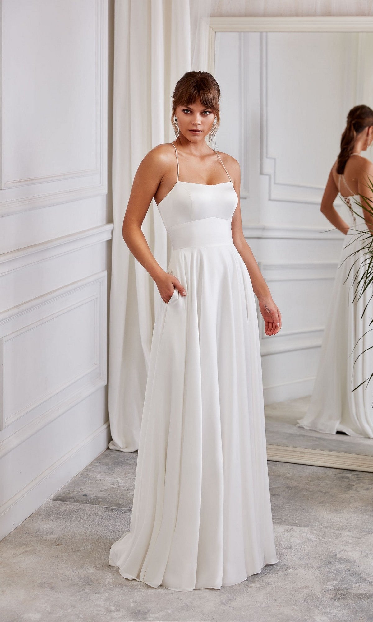 A woman stands indoors next to a large mirror and white curtains wearing the Aerie Couture 2005 long white dress, featuring thin straps, pockets, and a sleeveless, floor-length design.