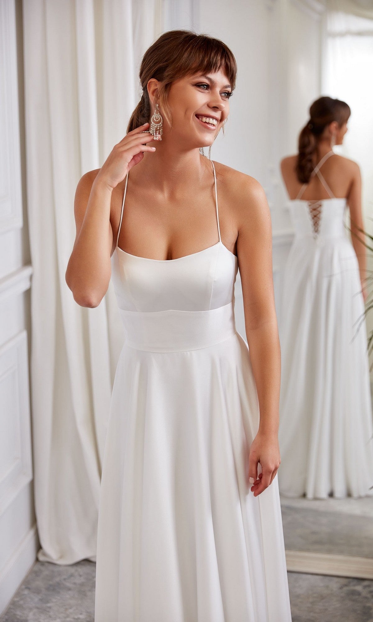A woman in the Aerie Couture 2005 long white dress with thin straps stands indoors, smiling and touching an earring, her reflection visible in a mirror behind her.