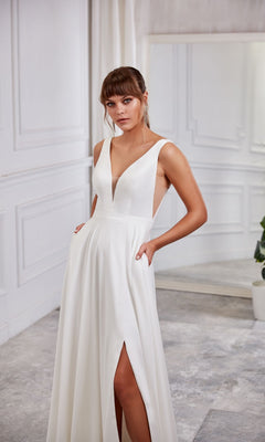 A woman stands in a white room with a mirror and potted plant, wearing the Aerie Couture 2007 Long White Dress—sleeveless, A-line, with a deep V-neckline and thigh-high slit.