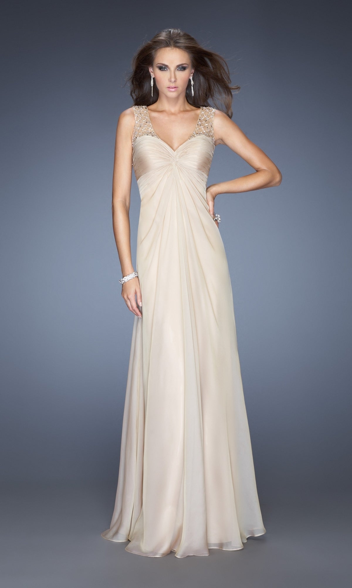 A woman models the La Femme Dress 20122, a floor-length, sleeveless beige chiffon gown with embellished straps and a draped bodice, set against a plain gray background.
