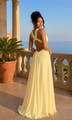 A woman in a long, light yellow La Femme Empire-Waist Ruched Chiffon Prom Dress 20134 stands on a terrace with columns, gazing out at the sea and palm trees beneath a clear sky.