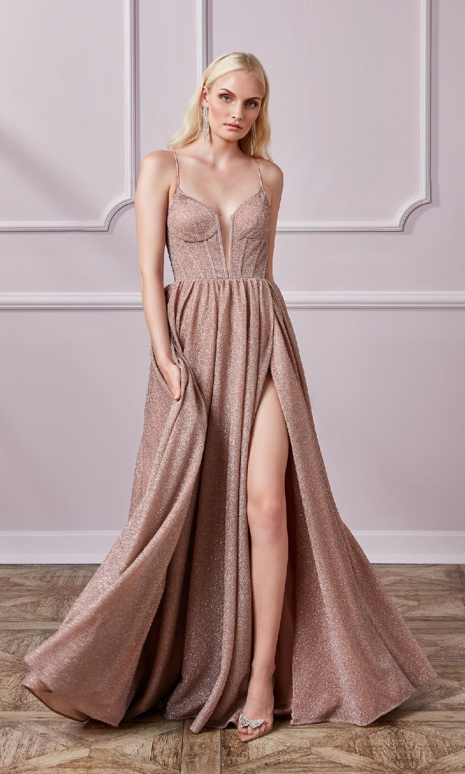 A woman models the Lace-Up Long Glitter Prom Dress: Aerie Couture 201, featuring shimmering blush pink fabric, spaghetti straps, a deep V neckline, and a high slit, standing against a pale pink paneled wall.