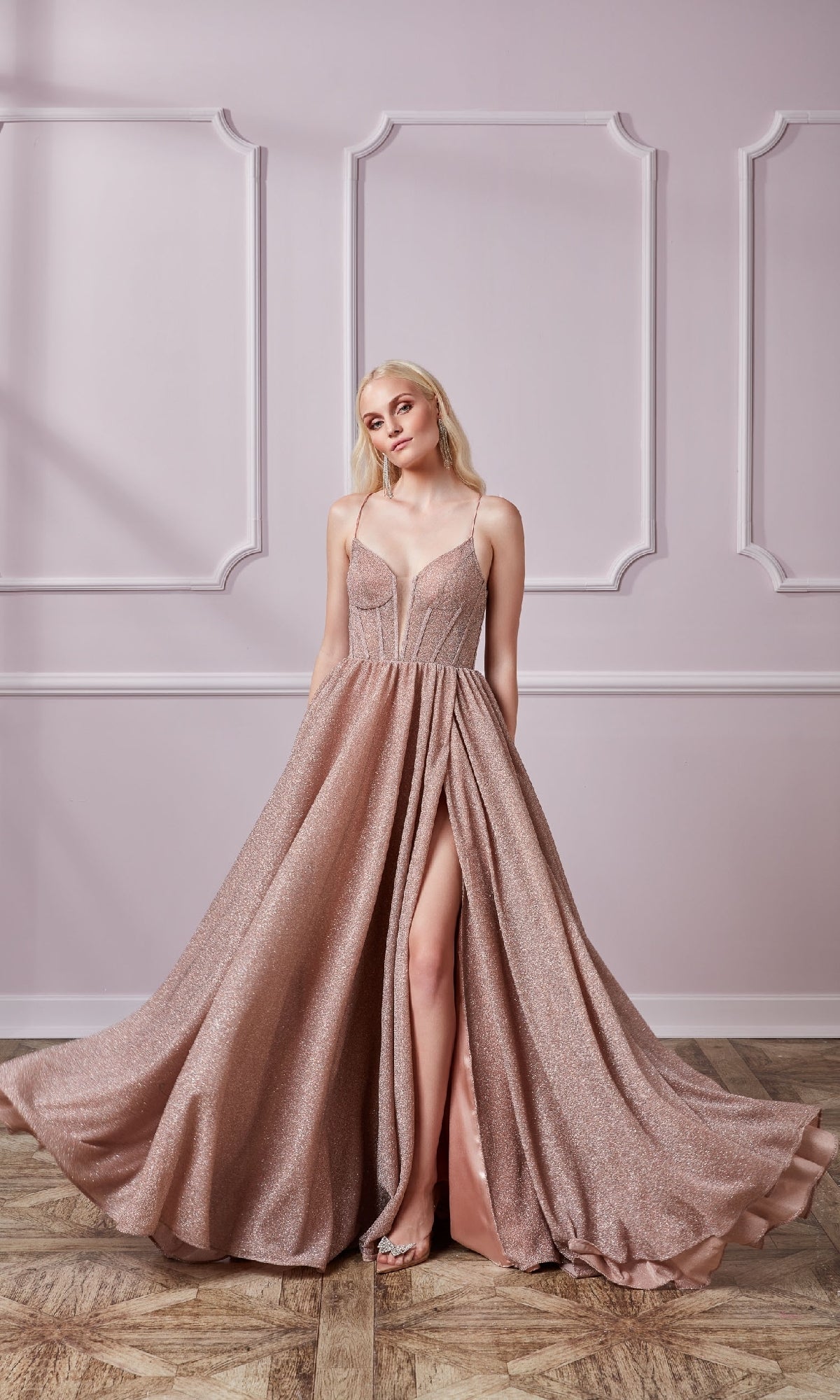A woman stands indoors wearing the Lace-Up Long Glitter Prom Dress: Aerie Couture 201, a shimmering blush pink A-line gown with spaghetti straps and a high slit, holding the sides of the formal dress.