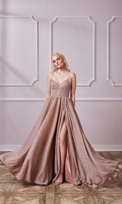 A woman stands indoors wearing the Lace-Up Long Glitter Prom Dress: Aerie Couture 201, a shimmering blush pink A-line gown with spaghetti straps and a high slit, holding the sides of the formal dress.