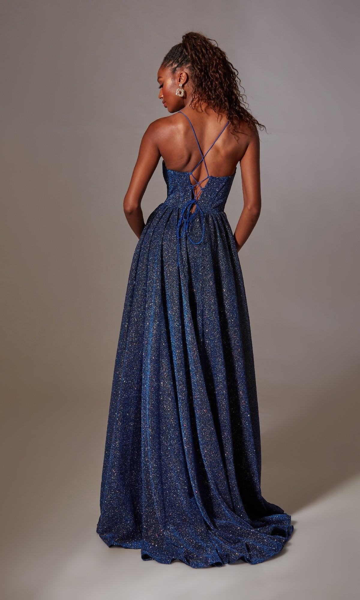 A woman faces away from the camera in the Lace-Up Long Glitter Prom Dress: Aerie Couture 201, a floor-length dark blue gown with thin cross-back straps and a sparkling, flowing skirt.