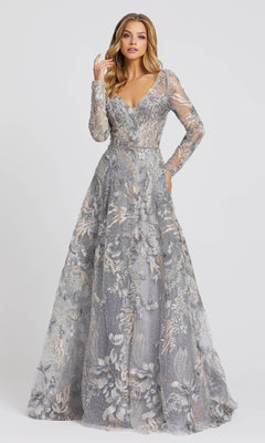 A woman wears the Mac Duggal 20214, a floor-length silver-gray formal dress with long sleeves, intricate floral embroidery, and a deep V-neckline, standing against a plain light background.
