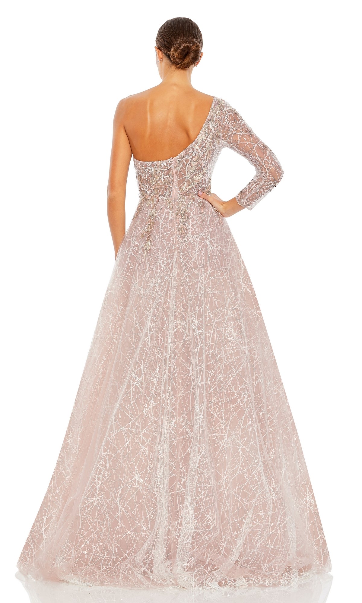 A woman stands facing away in the Long Formal Dress 20232 by Mac Duggal, a sparkling blush pink floor-length one shoulder ball gown with an asymmetrical design and her hair styled in a low bun.