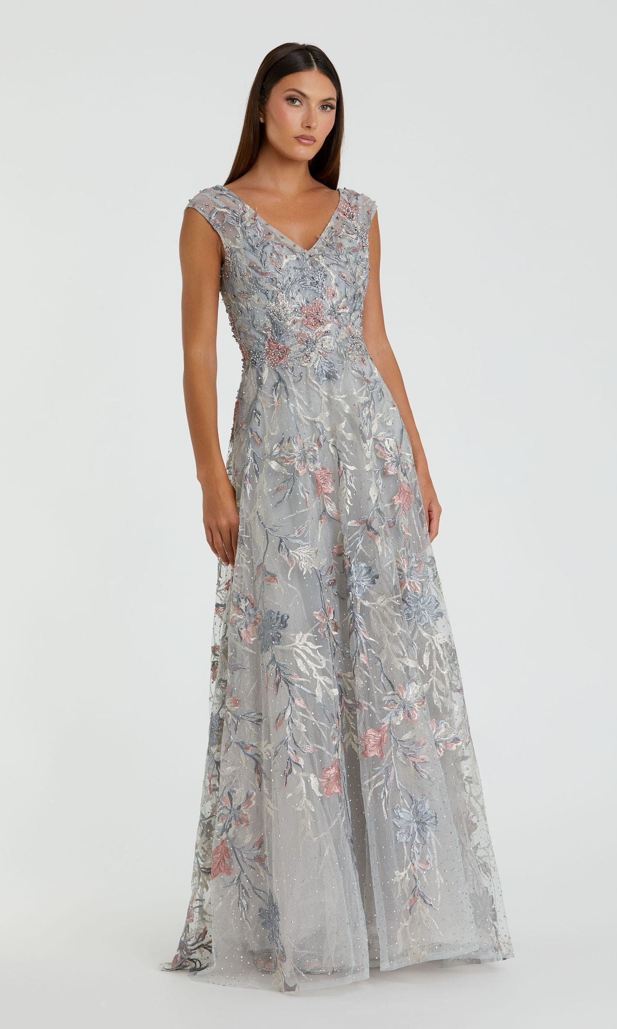 A woman wears the Mac Duggal 20263 Cap-Sleeve Long Print Prom Dress—a floor-length silver gown with cap sleeves, floral prints, and a V-neckline—standing against a plain light background.