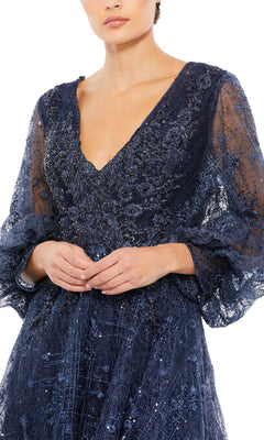 A woman wearing the Mac Duggal Long Formal Dress 20283, a navy blue sheer gown with sequins, embroidery, long sleeves, and a deep V-neckline, stands with her hands clasped in front of her.