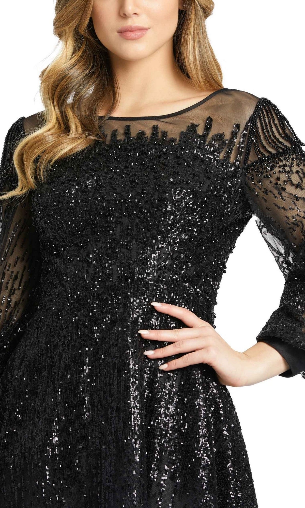 A woman poses with one hand on her waist in the Mac Duggal 20307 Long Sleeve Sequin Formal Gown, featuring a black design with a sheer neckline and sleeves.