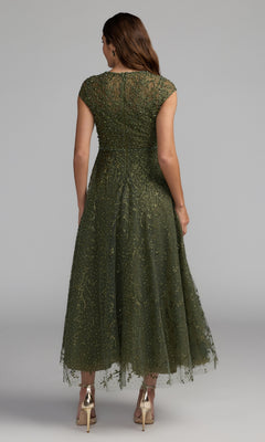 Woman standing with her back to the camera, wearing a lilac, tea-length Mac Duggal 20338 party dress with cap sleeves and beaded details, paired with gold high-heeled shoes.