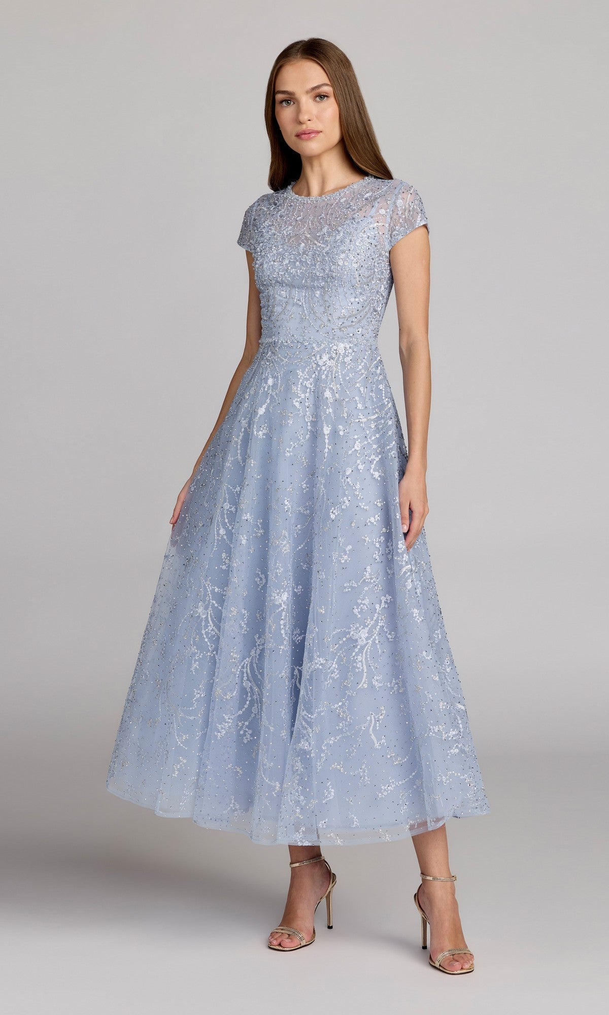 A woman stands wearing the Mac Duggal 20338 Tea Length Lilac Party Dress, featuring a fitted bodice, flared skirt, short sleeves, and delicate lace overlay. She completes her look with silver high-heeled sandals.