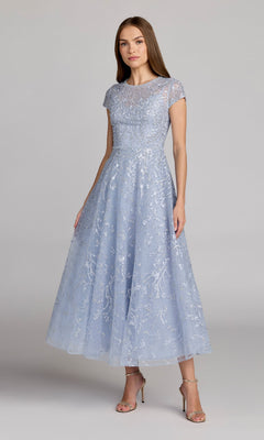 A woman stands wearing the Mac Duggal 20338 Tea Length Lilac Party Dress, featuring a fitted bodice, flared skirt, short sleeves, and delicate lace overlay. She completes her look with silver high-heeled sandals.