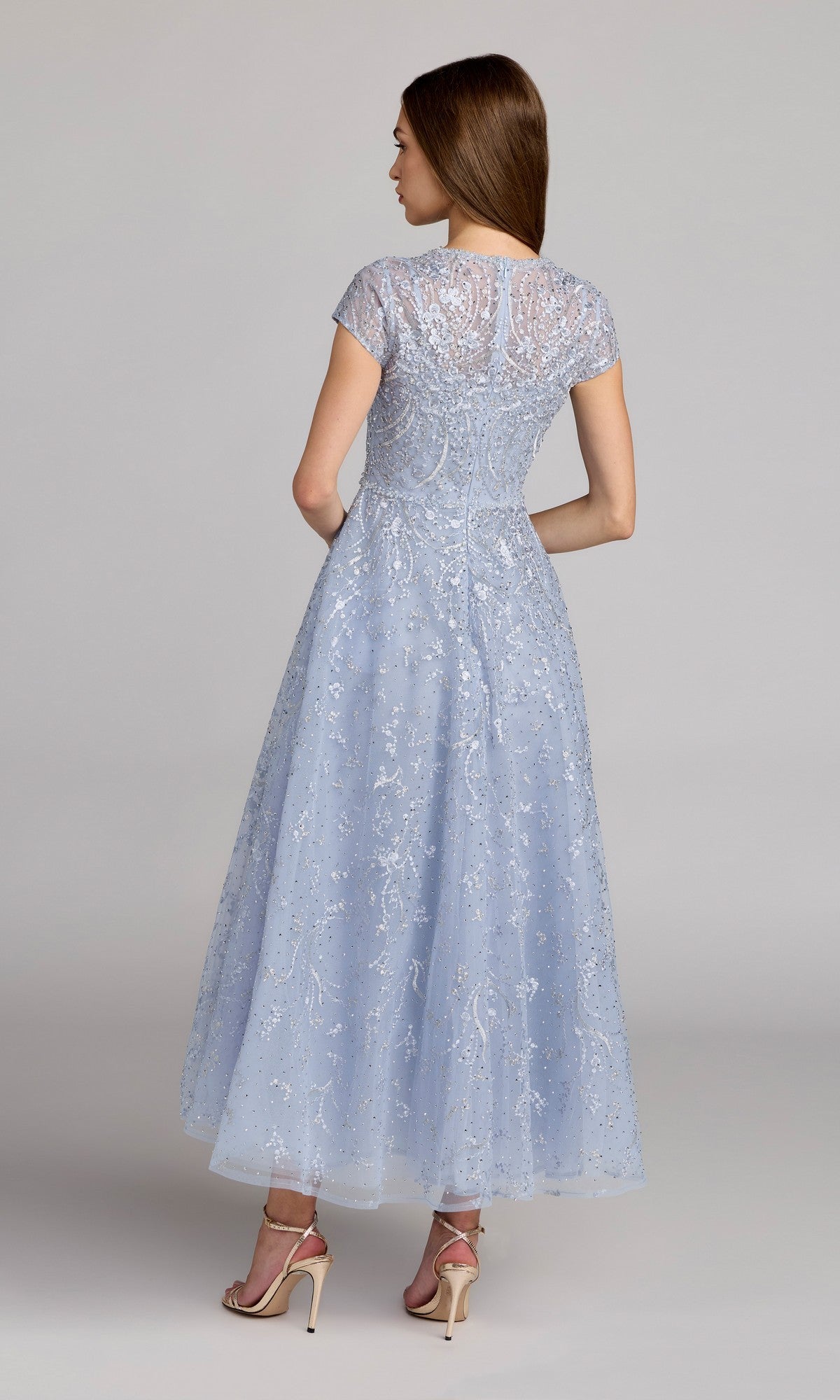 A woman faces away, wearing the Tea Length Lilac Party Dress: Mac Duggal 20338, featuring intricate beading and short sleeves, paired with nude high-heeled sandals against a plain background.