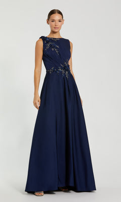 A woman stands against a plain light background wearing the Mac Duggal 20365— a sleeveless long formal navy blue dress with floral embroidery on the bodice.