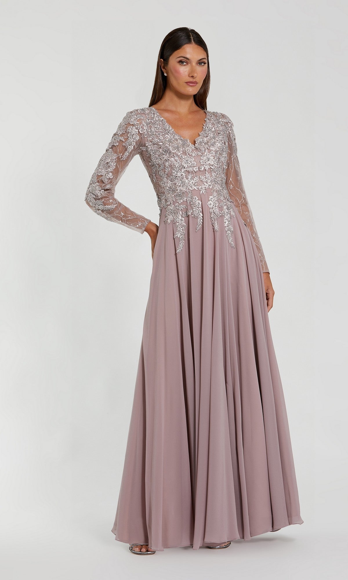 A woman stands against a plain light background wearing the Mac Duggal 20388, a mauve chiffon special occasion dress with sheer, lace-embroidered long sleeves.