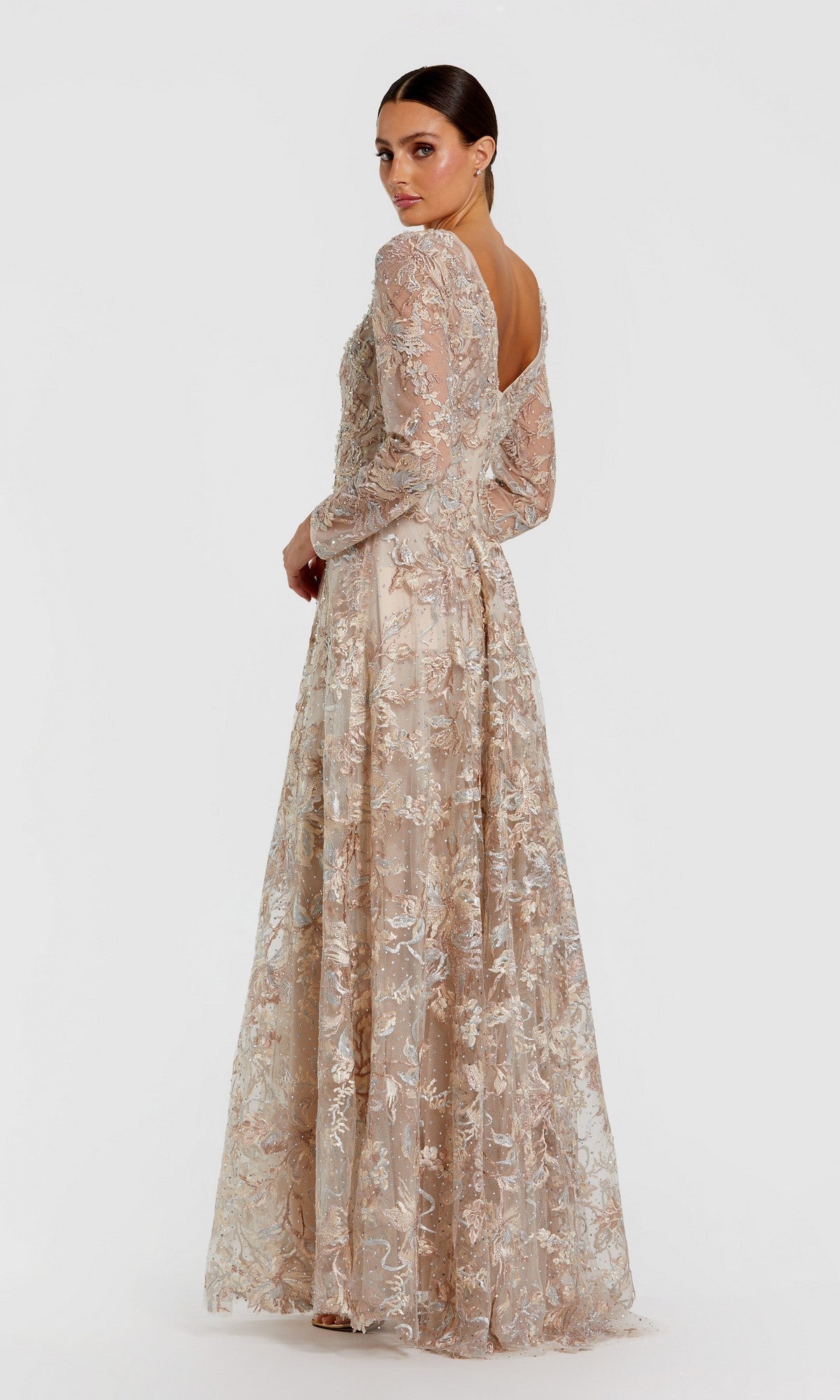 A woman models the Mac Duggal 20409 long formal dress, featuring light beige lace embroidery, long sleeves, and an open back, as she poses and looks over her shoulder against a plain white background.