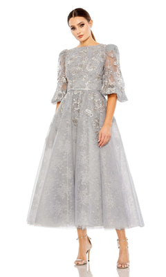 A woman models the Long Formal Dress 20418 by Mac Duggal, a light gray tea-length dress with puff sleeves and floral embroidery, paired with silver high-heeled sandals against a white backdrop.