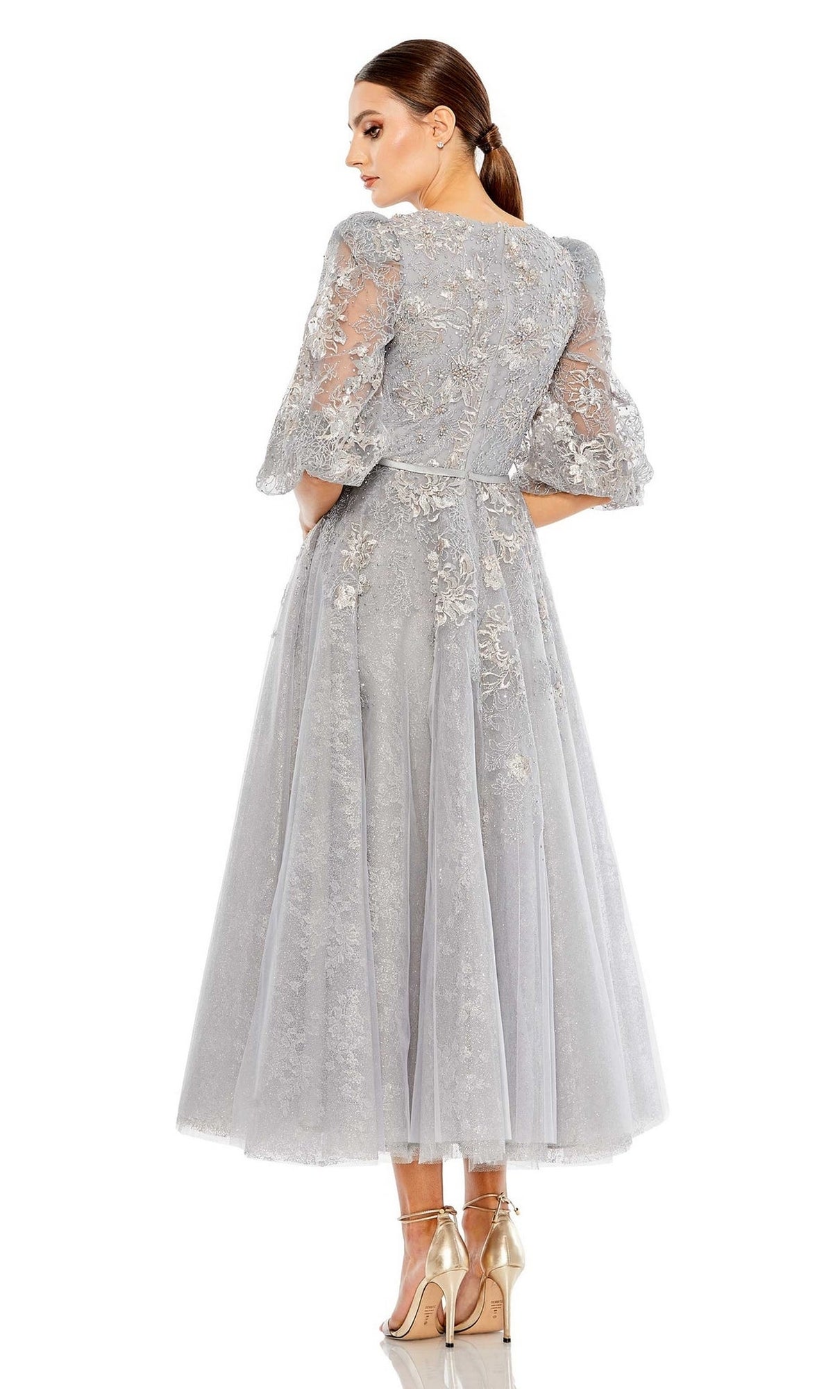 A woman faces away, wearing the Long Formal Dress 20418 by Mac Duggal—a tea-length light gray lace dress with puff sleeves and floral embroidery—paired with gold high heels.