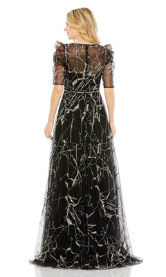 A woman stands facing away, wearing the Long Formal Dress 20428 by Mac Duggal—a black A-line gown with sheer sleeves, a beaded belt, and intricate silver branch-like patterns.