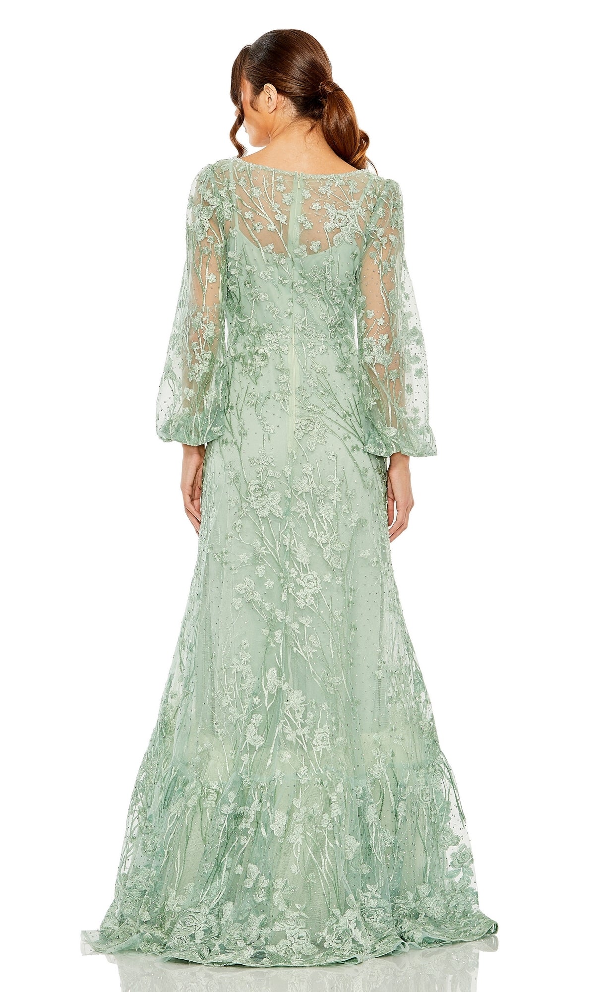 A woman in the Mac Duggal 20430 long sleeve formal dress, featuring sheer light green fabric with floral embroidery and a fitted waist, stands with her back to the camera.