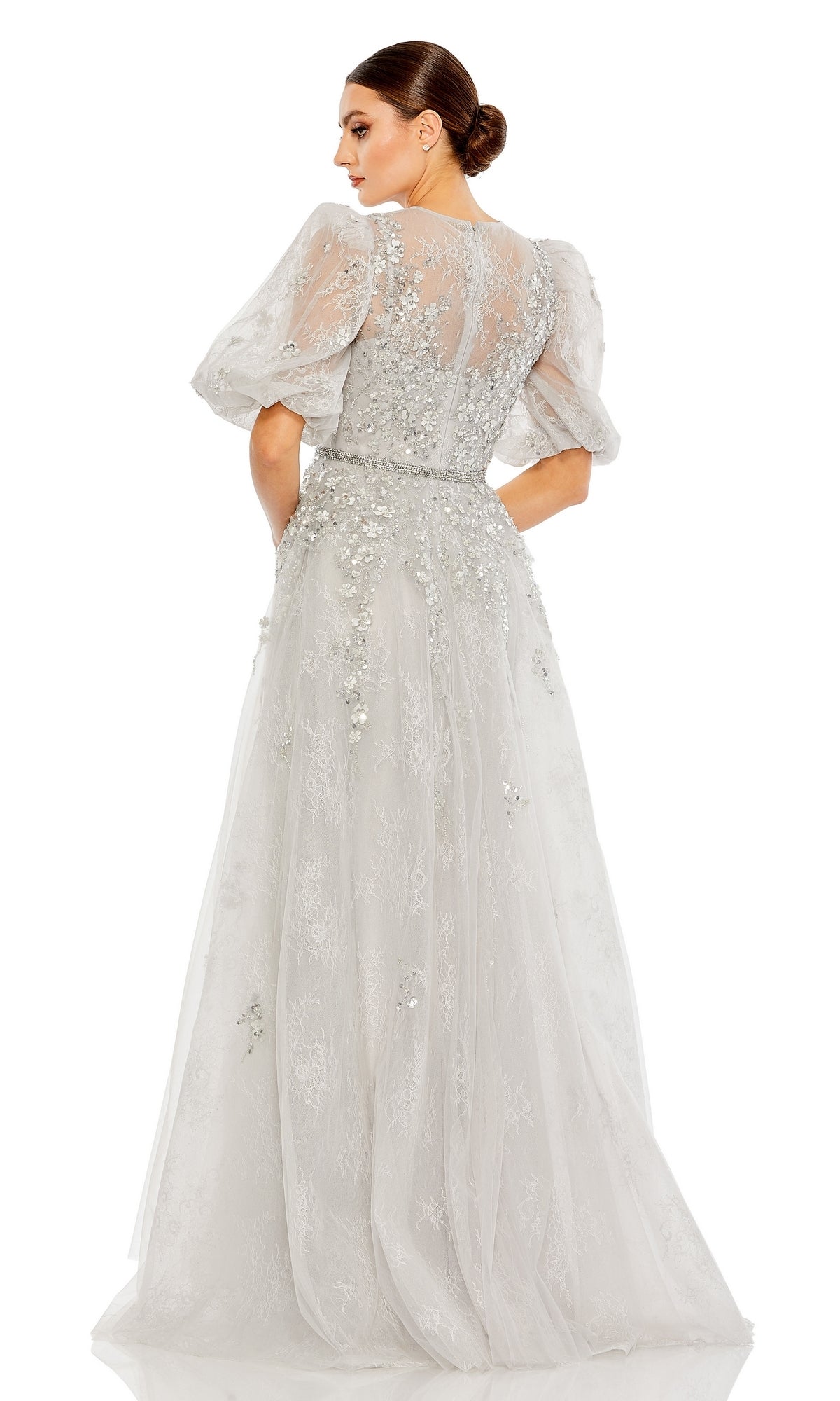 Woman wearing the Long Formal Dress 20445 by Mac Duggal, a floor-length platinum lace gown with sheer puff sleeves and floral embellishments, standing with her back to the camera.