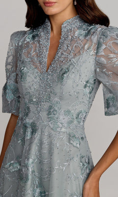 A woman wearing the Mac Duggal 20452 Short Sleeve Beaded Formal Gown in light blue with floral embroidery and a high collar, her face partially out of frame.