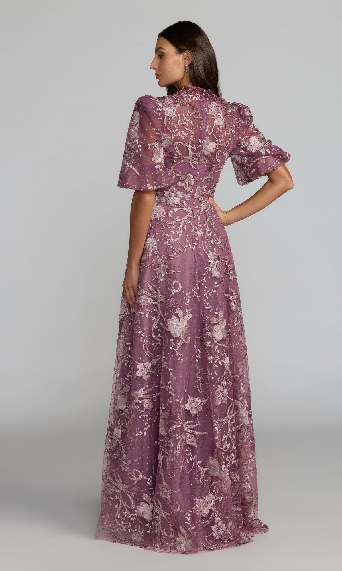 A woman stands in profile, modeling the Mac Duggal 20452 Short Sleeve Beaded Formal Gown— a floor-length mauve dress with floral patterns and puffed sleeves—against a plain, light-colored background.