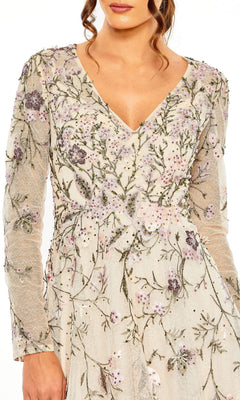 A woman wears the Mac Duggal 20480, a long-sleeve pink evening gown with a V-neck and delicate floral embroidery on sheer fabric.