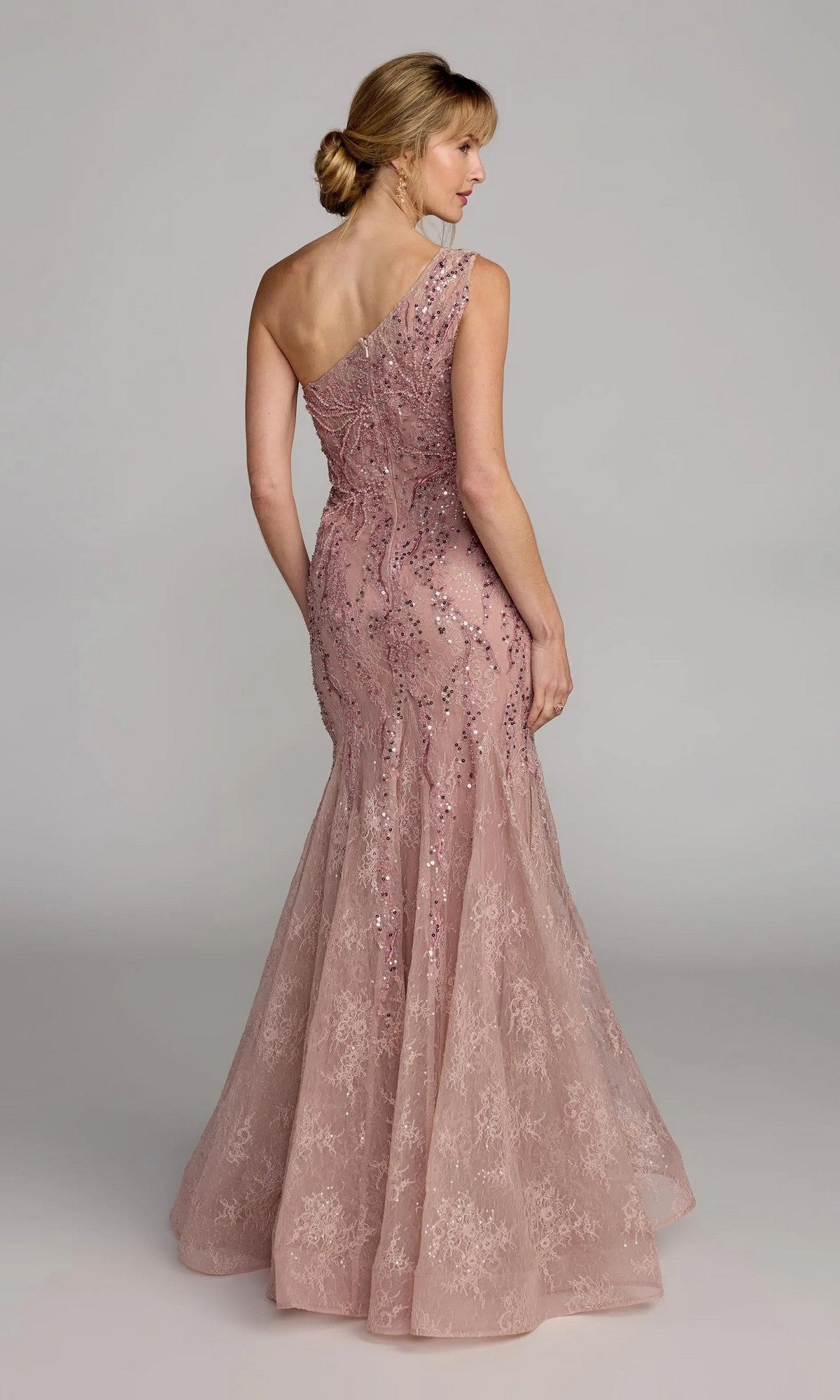 A woman stands with her back to the camera, wearing the Mac Duggal 20482—a fitted, one-shoulder blush pink mermaid gown featuring sequins and embellished lace—set against a plain background.