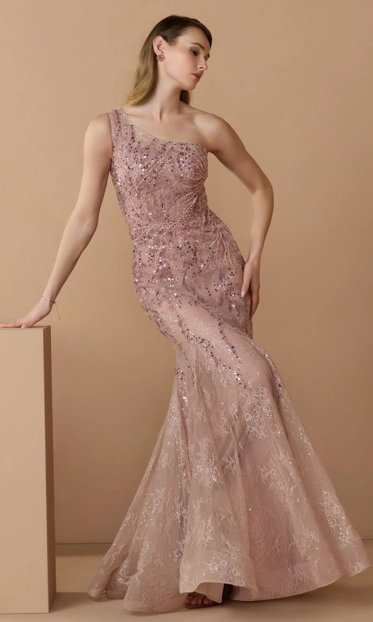 A woman models the Mac Duggal 20482 long formal dress in rose, a one-shoulder, floor-length mermaid gown with sequin and embellished lace details, set against a beige background.