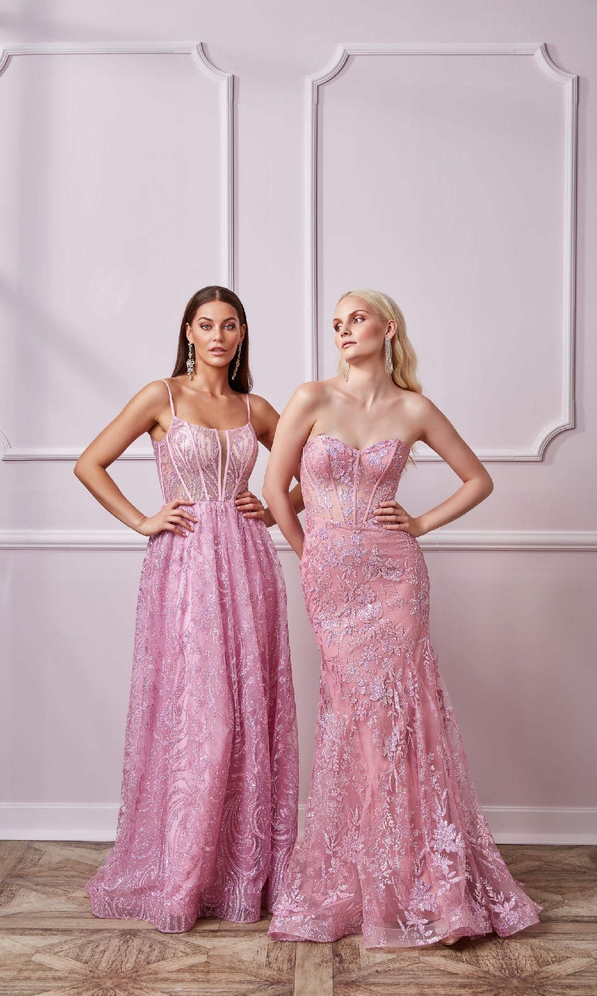 Two women stand side by side in a room with light-colored paneled walls and wooden floors, wearing the Sparkly Long Mermaid Prom Dress: Aerie Couture 204—floor-length pink lace gowns with corset bodices and intricate detailing.