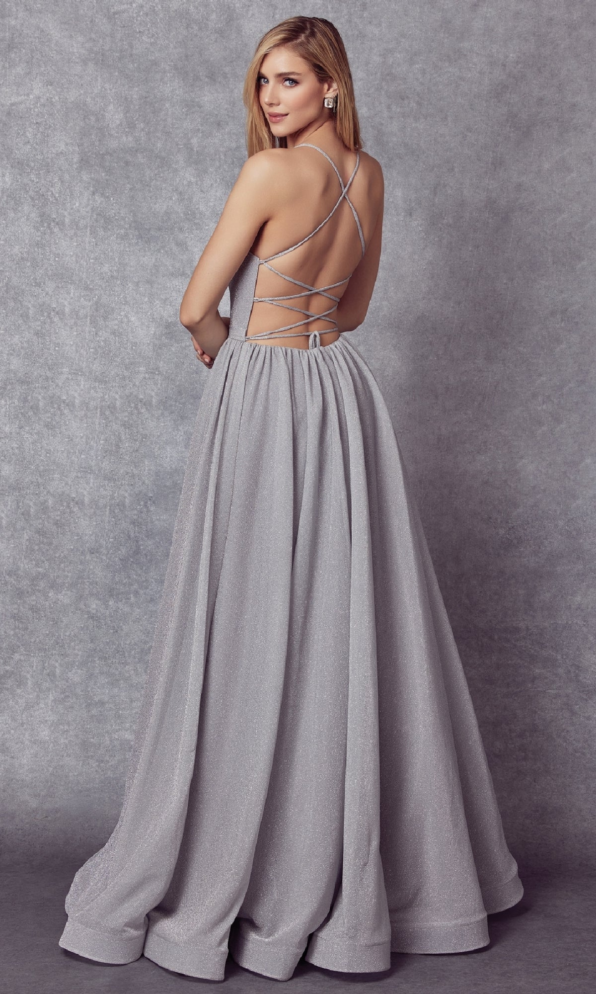 A woman models the Juliet 204 Long Prom Dress—silver, floor-length, with a shimmer finish and crisscross open back—in front of a gray textured backdrop, standing slightly turned from the camera.