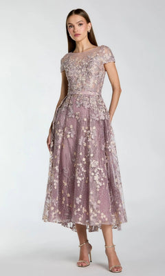 A woman wearing the Mac Duggal 20502 tea-length party dress in vintage lilac, featuring floral embroidery, delicate applique, short sleeves, and a sheer overlay, stands in neutral heels against a plain background.