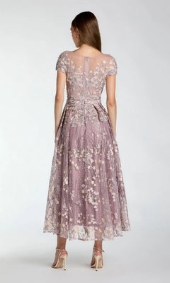 A woman stands facing away, wearing the Mac Duggal 20502 tea-length party dress in mauve with delicate lace floral appliqué and short sleeves, paired with high-heeled sandals against a plain light background—evoking the refined elegance characteristic of Mac Duggal’s lilac-inspired collections.