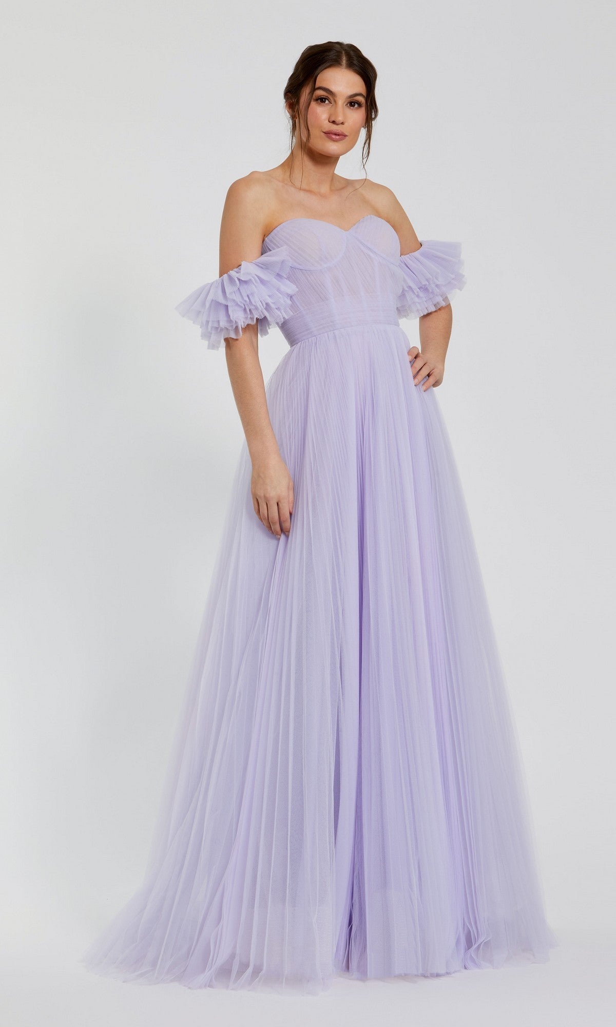 A woman stands against a plain light background in the Mac Duggal 20517 long formal dress, an off-the-shoulder lilac gown with pleated tulle and ruffled sleeves.