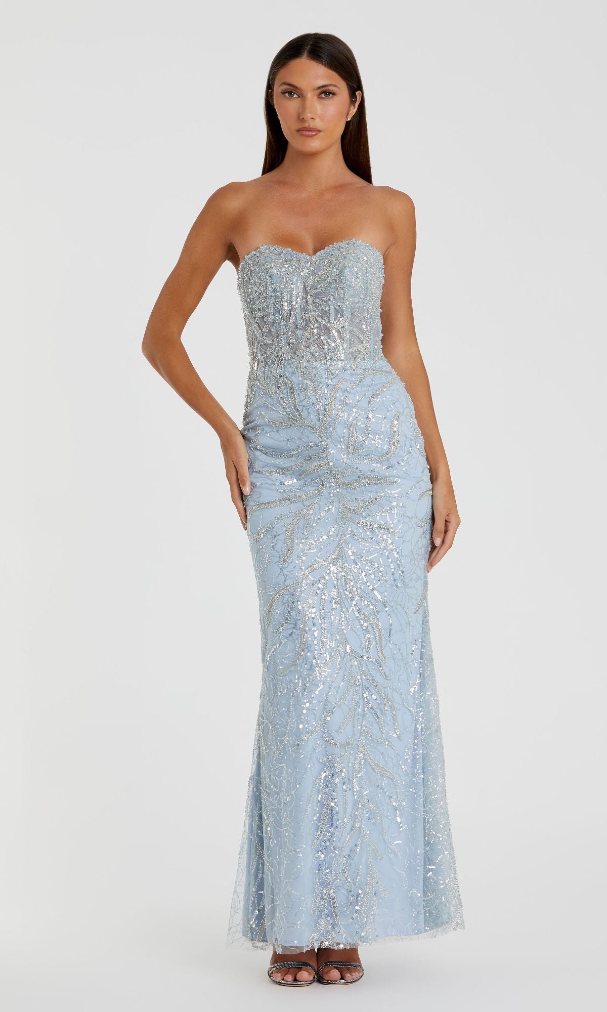 A woman models the Mac Duggal 20544 Strapless Long Beaded Prom Dress in light blue with silver sequins, standing before a plain white background.