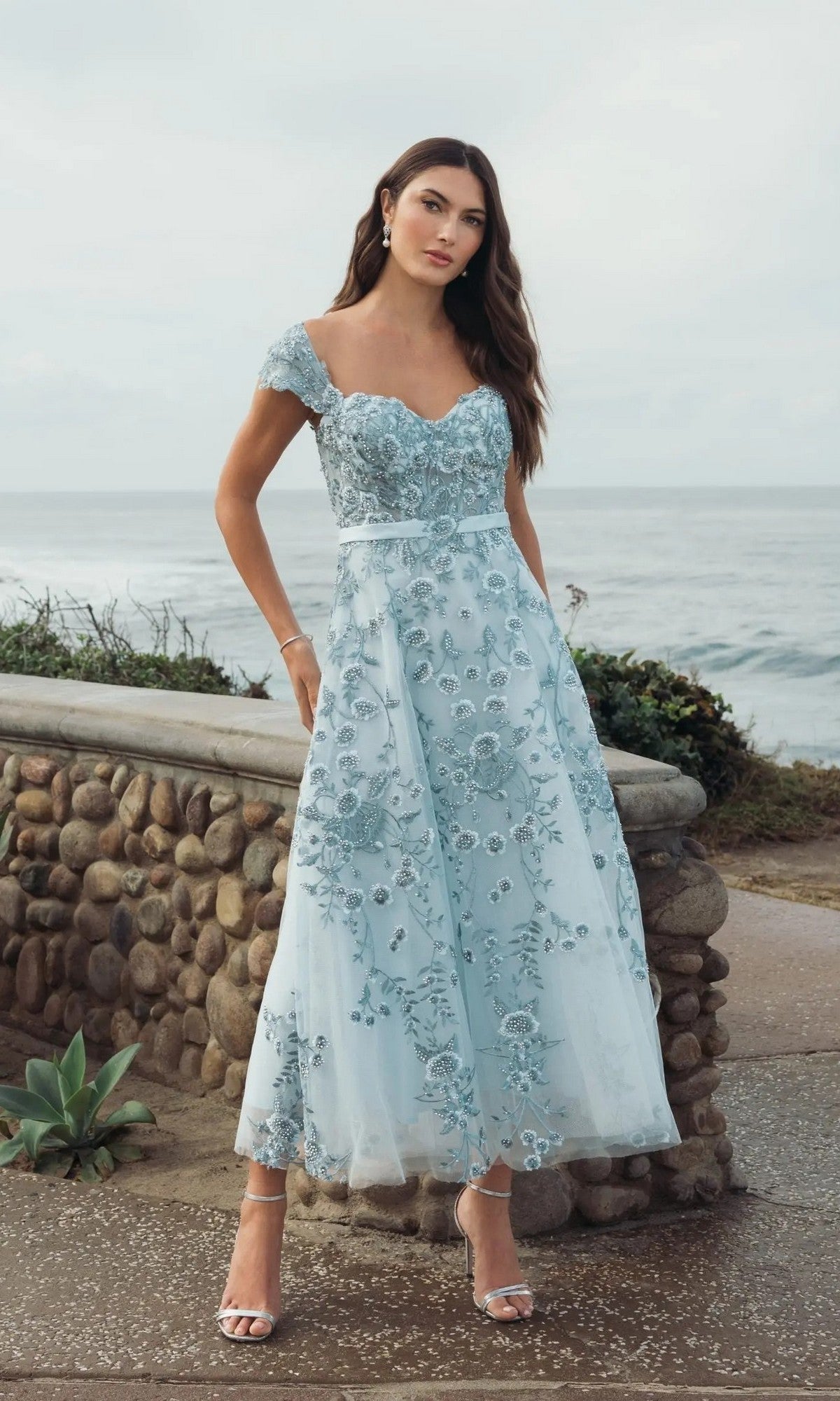 A woman stands outdoors by a stone wall with the ocean behind her, wearing the Cap-Sleeve Nude Midi Party Dress: Mac Duggal 20582, featuring elegant floral embroidery.