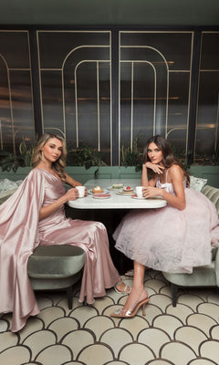 Two women wearing Short Homecoming Dress 20583 by Mac Duggal in rose pink sit at a round table in a chic café, enjoying coffee and pastries together.