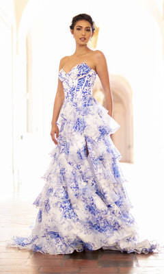 A woman stands in a bright corridor wearing the Amelia Couture 2060 Strapless Print Prom Gown, featuring a corset-style bodice and ruffled ball gown skirt in a blue and white floral pattern.