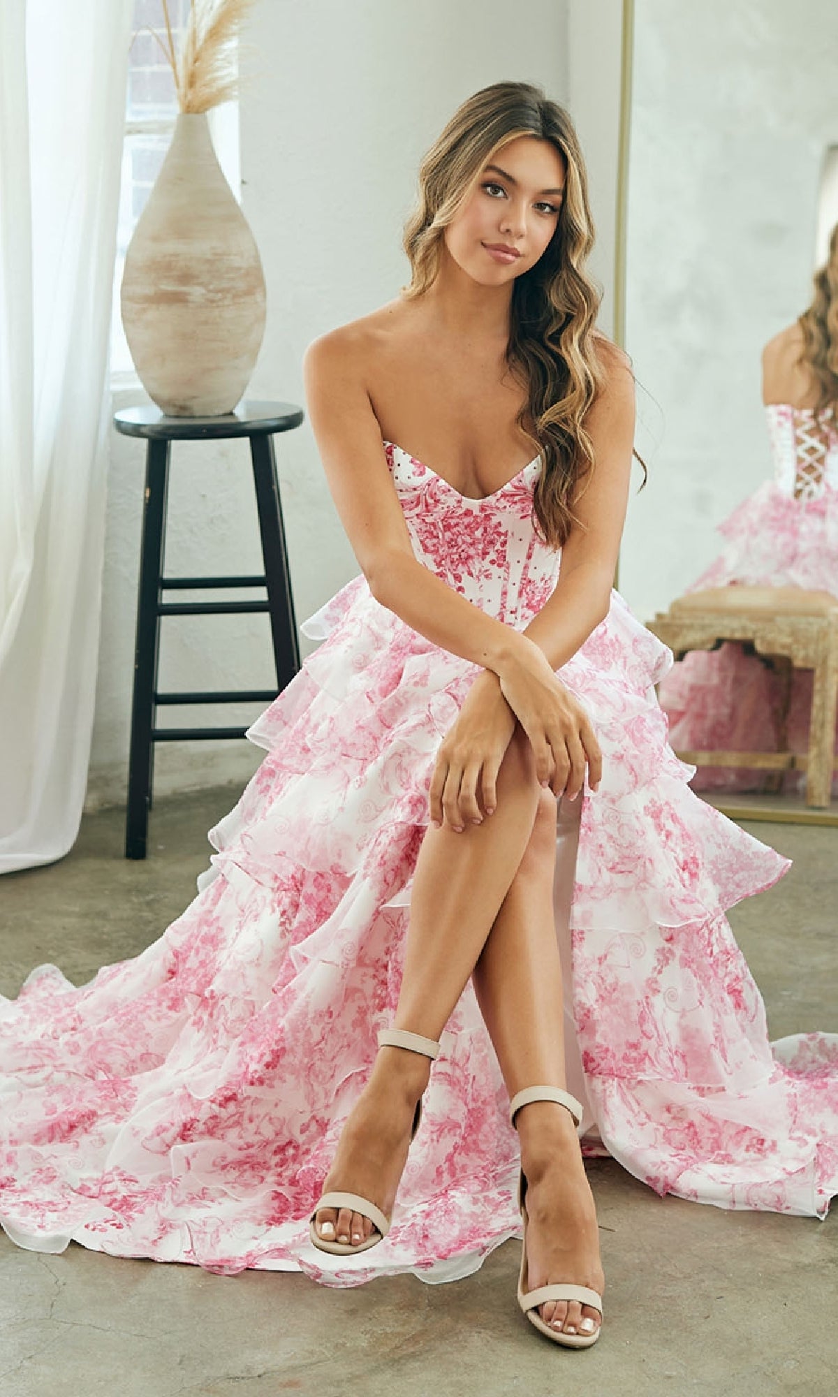 A woman in the pink Strapless Print Prom Gown by Amelia Couture 2060 sits on a stool with legs crossed, white heels on, in a softly lit room with neutral decor. The gown’s corset-style bodice highlights her silhouette.