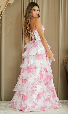 Woman wearing the Strapless Print Prom Gown: Amelia Couture 2060, a strapless long floral dress with a corset-style bodice and pink and white tiered layers, stands in front of beige curtains.