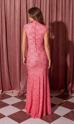 A woman stands on a checkered floor facing away, wearing the Eureka 2061 Long Lace Formal Dress—a sleeveless, floor-length pink lace gown with a high neck—in front of pink curtains.
