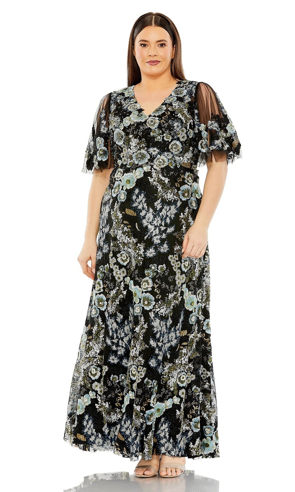 A woman wearing the Long Plus-Size Prom Dress 20630 by Mac Duggal—featuring black floral embroidery, sheer short sleeves, and paired with beige heels—stands facing forward against a white background.