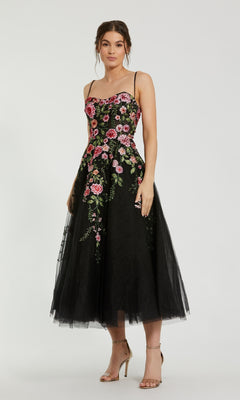 A woman wearing the Mac Duggal 20651 Floral Embroidered A-Line Midi Dress, featuring black fabric, thin straps, and colorful floral embroidery, stands against a plain background.