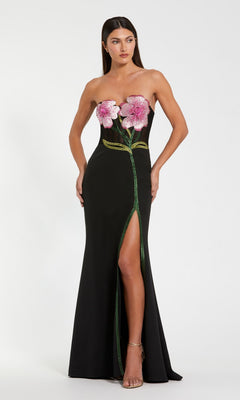 A woman wears the Mac Duggal 20682 long formal dress in black, featuring a strapless crepe design with pink flower appliqués on the bodice, a green stem down the front, and a high leg slit—ideal for special occasions.