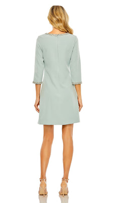 A woman faces away, wearing the Mac Duggal 20697 short wedding guest dress in seafoam green with three-quarter sleeves and embellished trim, styled with nude heels.